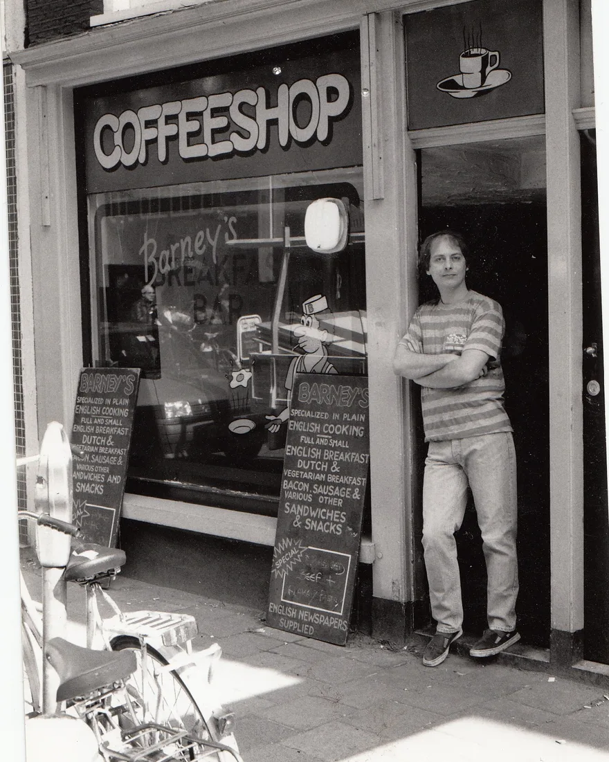 Nick outside Barney’s Breakfast Bar in 1986, arms folded, beside boards advertising breakfast and English newspapers.
